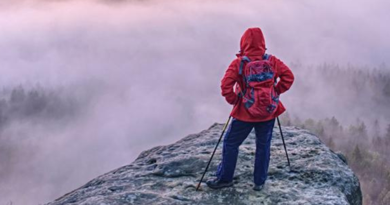 Person standing on a rocky overlook above a fog-covered landscape, looking out across a complex and uncertain horizon.